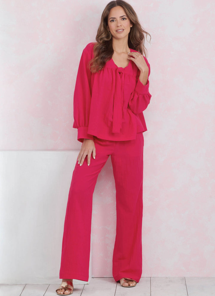 In front of a delicate pink backdrop, a woman is dressed in Super Cheap Fabrics' Simplicity 9928 ensemble, featuring a vibrant pink lounge blouse and coordinating wide-leg trousers. Her long brown hair cascades down as she softly smiles, her hands resting at her side. Completing the look are bronze open-toe sandals and a spacious tote.