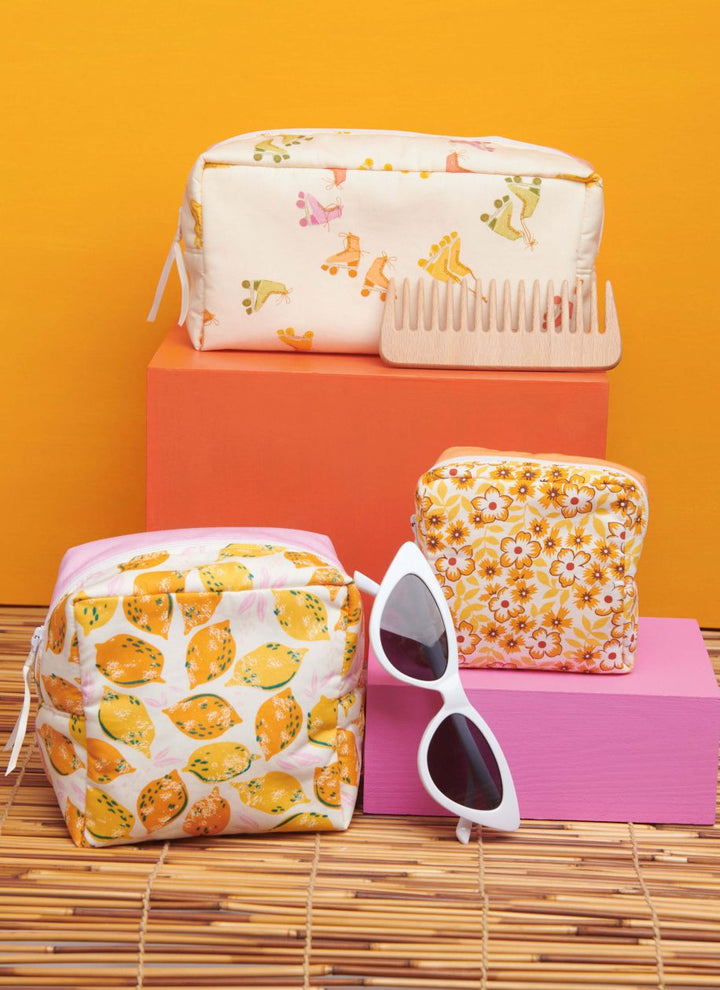 A vibrant display of toiletry bags featuring floral and fruit designs from Super Cheap Fabrics' Pattern - Simplicity - 9937 collection is set against an orange backdrop, reminiscent of the allure of empire waist tops. The scene also includes a wooden comb and white cat-eye sunglasses, with pink and orange blocks providing varying heights.