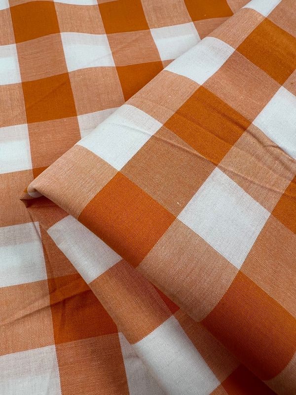 Close-up of the Cotton Gingham XL fabric from Super Cheap Fabrics, featuring an orange and white checkered pattern. Made from natural cotton fibers, this 112cm wide material is slightly wrinkled and layered, showcasing its bold geometric design that’s perfect for home decor.