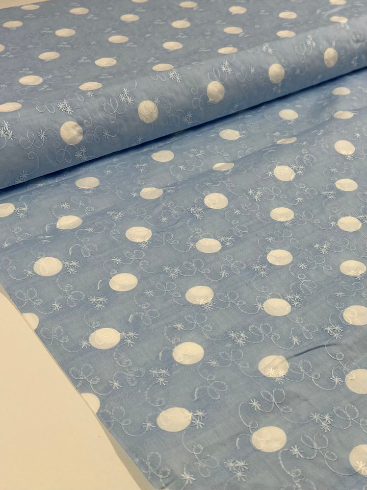 The "Embroidered Printed Cotton - Baby Blue - 112cm" from Super Cheap Fabrics showcases a delightful design of white polka dots adorned with intricate embroidered swirls and snowflakes, making it an ideal choice for crafting charming clothing and household items.