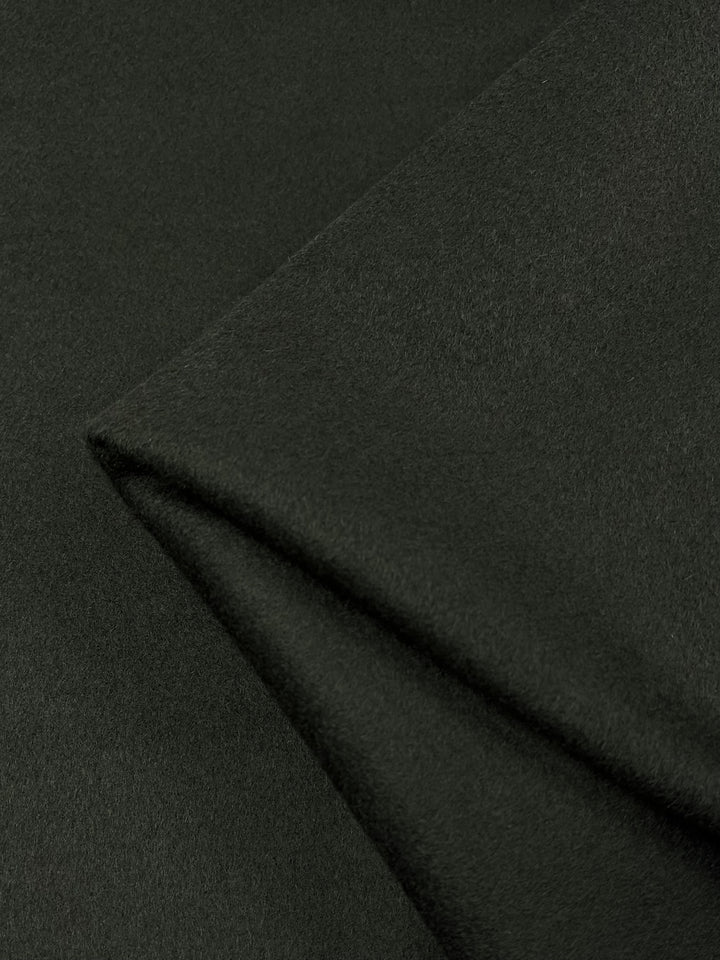 Close-up image of dark green Wool Cashmere - Forest Night - 150cm by Super Cheap Fabrics with a soft and slightly fuzzy texture. The fabric is folded, showcasing its medium to heavy weight and smooth surface. The color appears consistent throughout the material, reminiscent of a forest night fabric.