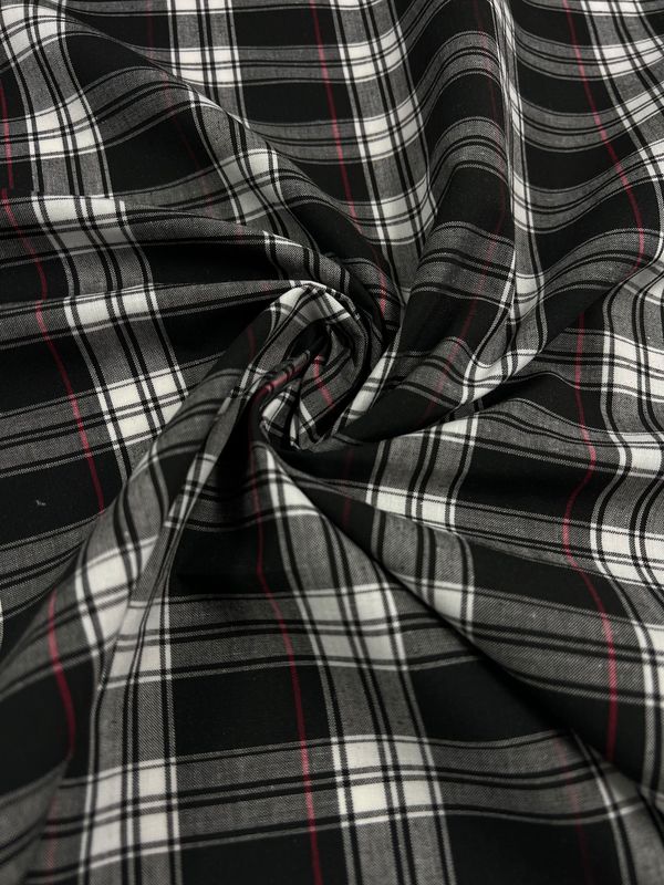 A detailed view of the Shirting - Douglas fabric from Super Cheap Fabrics reveals its lightweight blend with a striking black, white, and red plaid pattern. The polyester cotton material gracefully drapes in soft folds originating from the center, highlighting the intersecting lines and vivid color contrasts, making it perfect for versatile clothing applications.