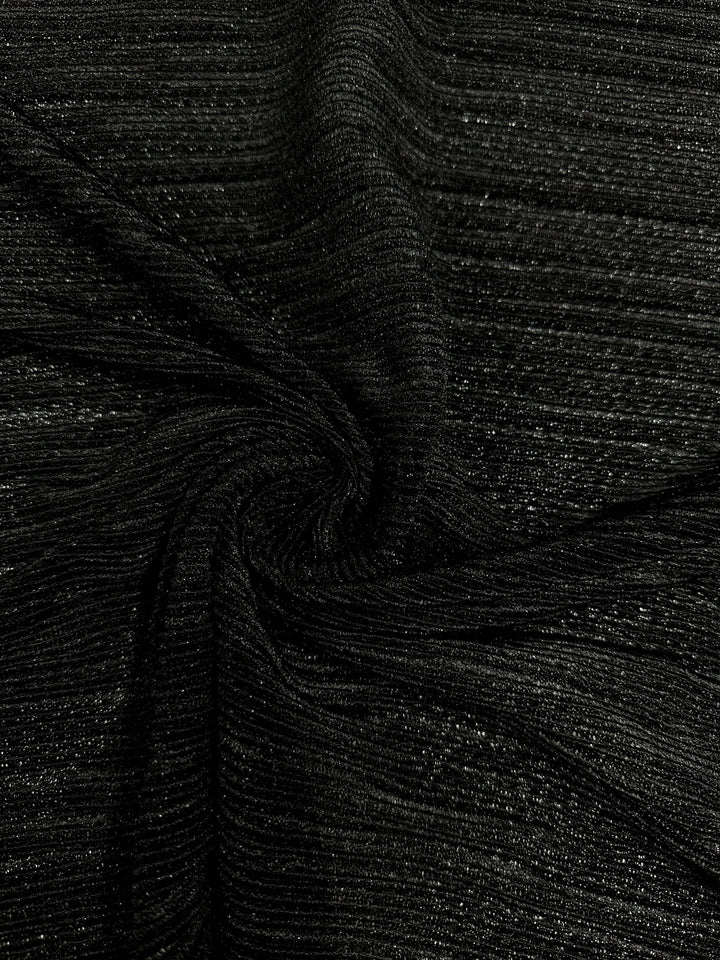 Close-up of the *Metallic Knit - Black - 150cm* fabric by **Super Cheap Fabrics**, characterized by its black color interwoven with subtle, glittery threads running horizontally. The fabric is gathered and slightly twisted in the center, creating soft folds and a swirled pattern. Perfect for chic dresses, this medium to heavyweight material appears soft and stretchy with a shiny finish.