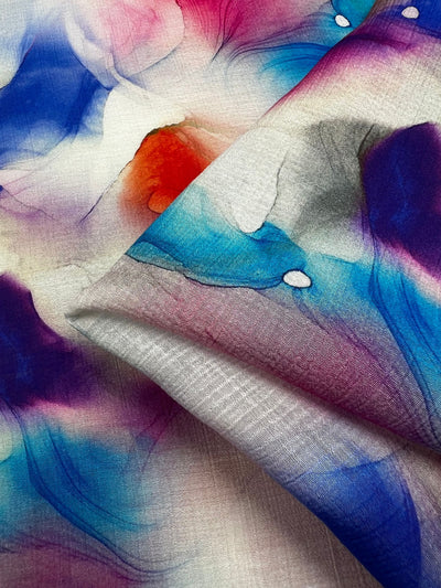 Here's a revised version of the sentence using the given product data:

A close-up of Super Cheap Fabrics' Designer Cotton - Crysta - 150cm showcases its vivid watercolor design, blending shades of blue, purple, pink, and orange. The abstract pattern flows across the material, evoking images of ink blots or fluid art, making it ideal for luxury sewing projects.