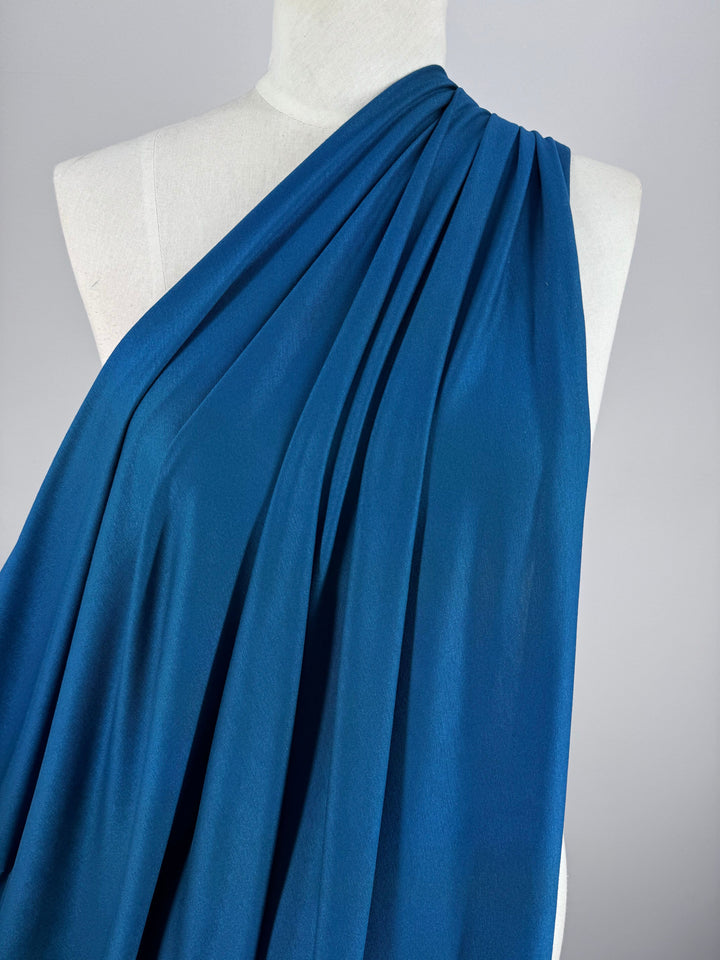 A mannequin draped in Super Cheap Fabrics' Cotton Lycra - Lyons Blue showcases a stretchy, single-shoulder pleated design for an elegant look. The plain light gray background enhances the movement and allure of this 150cm wide fabric.