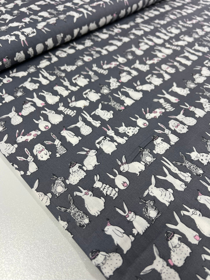 A fabric from Super Cheap Fabrics, named "Printed Cotton - Bunny Mayhem - 120cm," showcases a repeated design of white rabbits against a dark gray backdrop. The rabbits, made from natural cotton fibers, display various poses with pink accents scattered throughout. This consistent pattern spans the entire fabric, making it ideal for both clothing and home decor projects.