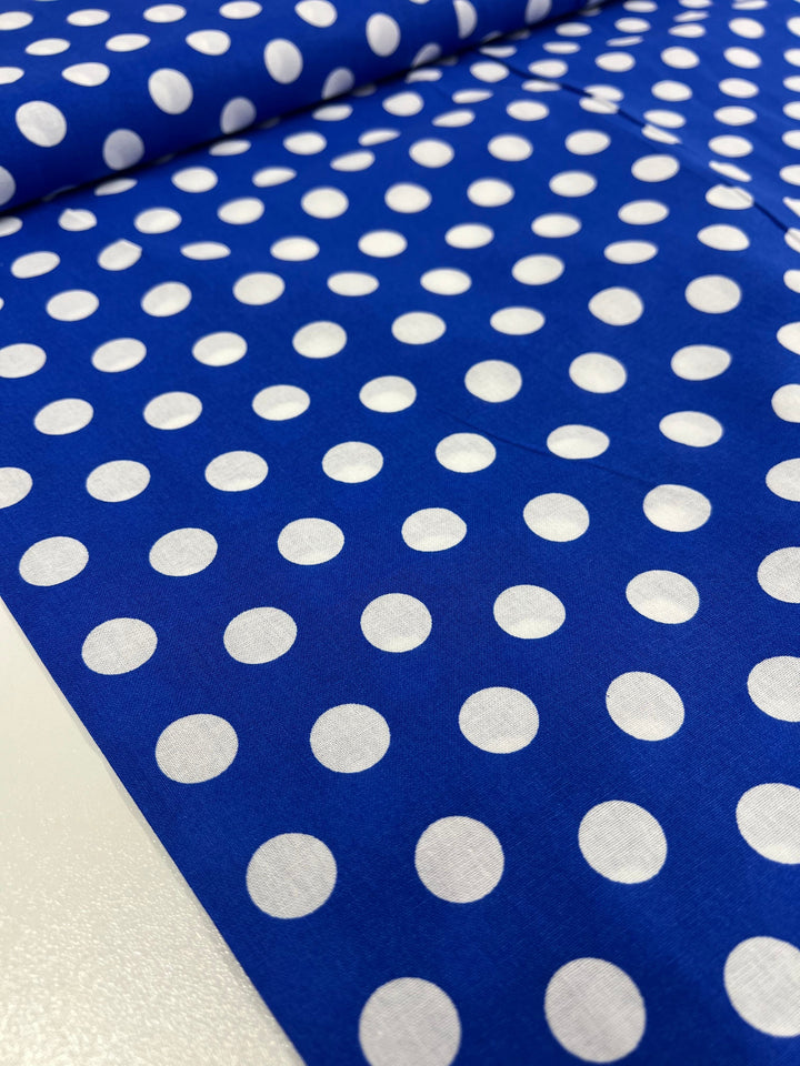 A roll of Linen Blend - Dazzling Blue - 112cm from Super Cheap Fabrics lays partially unrolled on a smooth surface, showcasing its lightweight and versatile qualities with a white polka dot pattern.