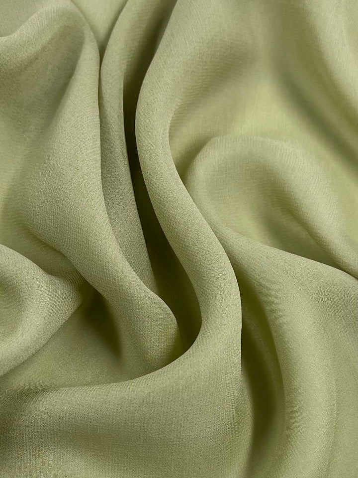 A close-up of "Silk Georgette - Celery - 135cm" by Super Cheap Fabrics reveals its light green hue with soft, flowing folds. The texture appears smooth, and this lightweight fabric forms gentle curves and creases, making it perfect for creating delicate and elegant spring and summer outfits.