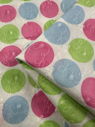 Introducing the "Embroidered Printed Cotton - Candy - 112cm" by Super Cheap Fabrics, this exquisite material showcases a vibrant pattern of pink, blue, and green circles on a white background. Enhanced with delicate white embroidery featuring swirls and stars, it is expertly crafted from natural cotton fibers. Ideal for both clothing and household projects, the fabric is beautifully displayed in a folded close-up view.