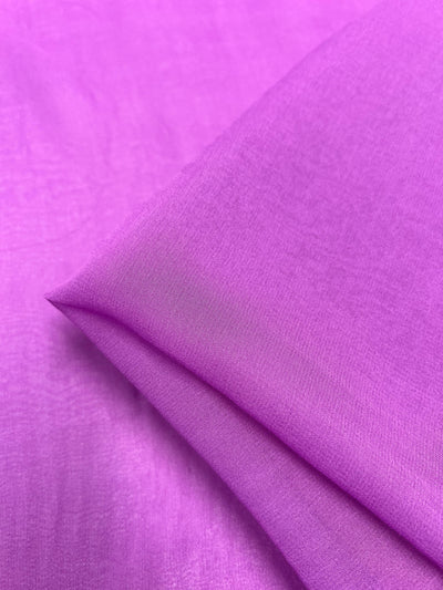 A close-up of the Silk Georgette - Iris - 135cm fabric from Super Cheap Fabrics showcases its vibrant purple hue and smooth, slightly translucent texture. One corner of this lightweight fabric is folded over, creating a gentle crease that adds depth to the image—perfect for spring and summer outfits.