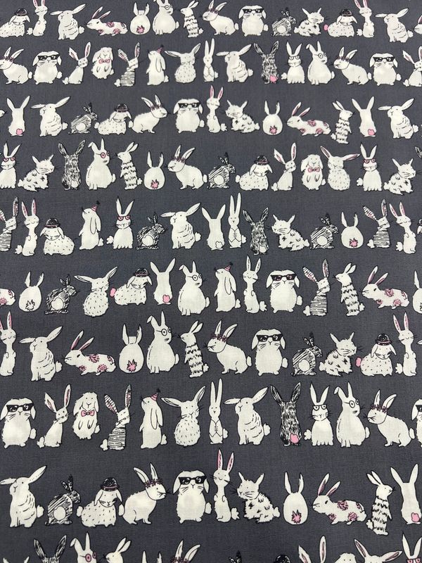 The "Bunny Mayhem" printed cotton fabric from Super Cheap Fabrics showcases an array of white bunnies on a dark gray background, with some donning glasses or accessories. This charming and whimsical design is perfect for crafting clothing and household items using natural cotton fibers. The fabric measures 120cm in width.