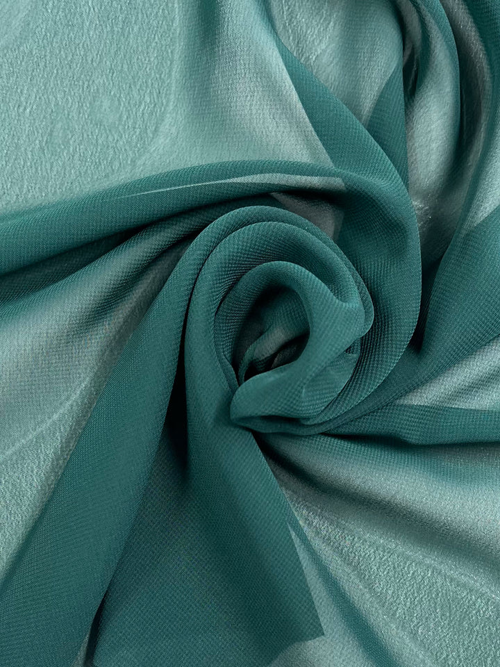 A close-up of Hi-Multi Chiffon - Dark Green - 150cm by Super Cheap Fabrics. The lightweight, sheer material is gathered and twisted into a loose spiral, showcasing its translucent and flowing texture. The background appears to be the same 100% polyester fabric, creating a cohesive and elegant appearance.