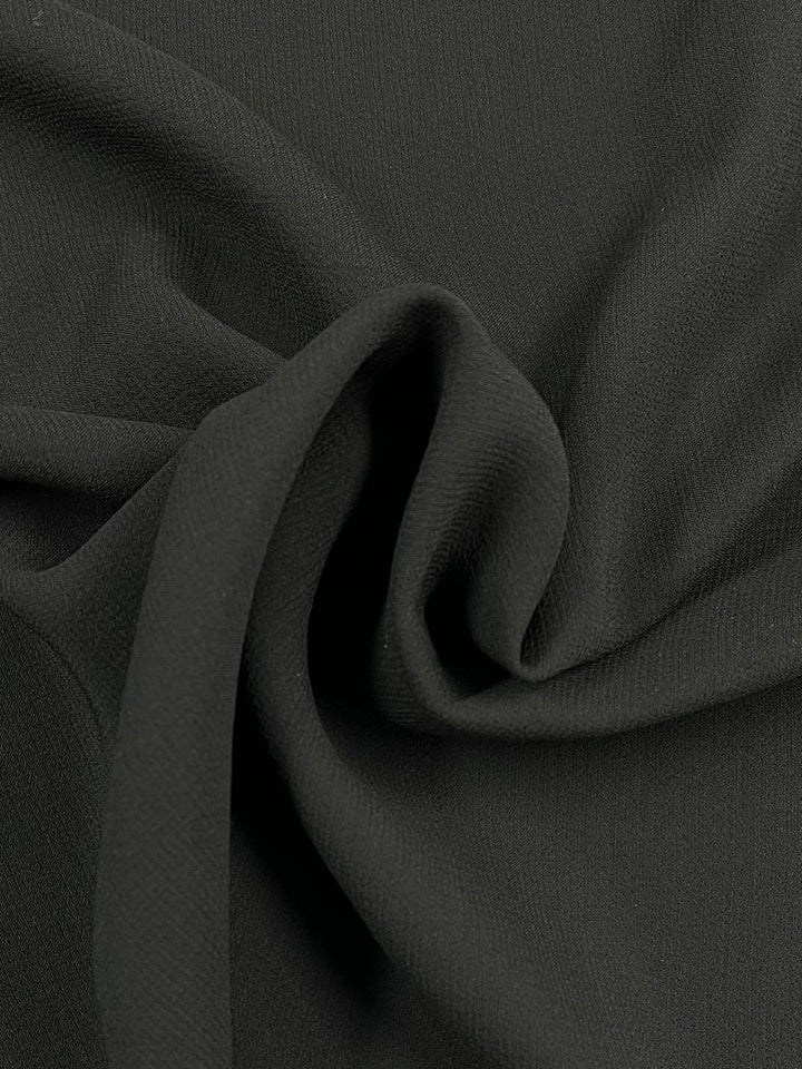 Close-up of Super Cheap Fabrics' Plain Crepe - Black (140cm), featuring a textured surface with gentle folds and a twisted center. This medium weight fabric offers an elegant drape and subtle sheen, creating soft shadows and highlights.