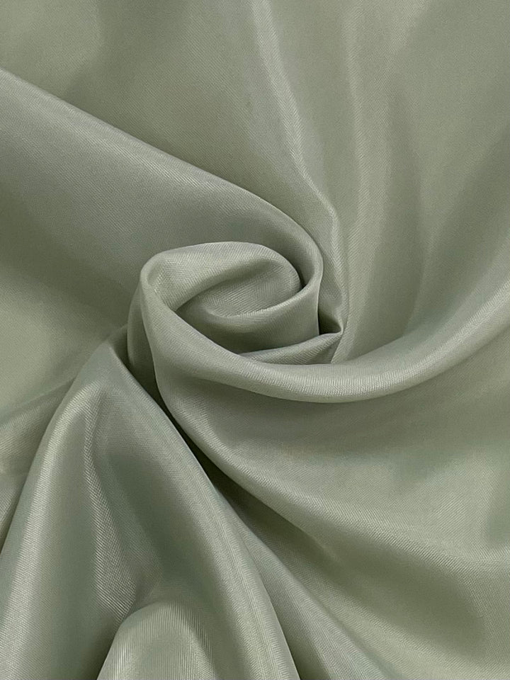 A close-up of a soft, light green satin fabric, gently wrinkled and gathered in swirling folds, creating a smooth and lustrous texture. The Lining - Ambrosia - 120cm by Super Cheap Fabrics catches light like ambrosia, highlighting its sheen and delicate draping.