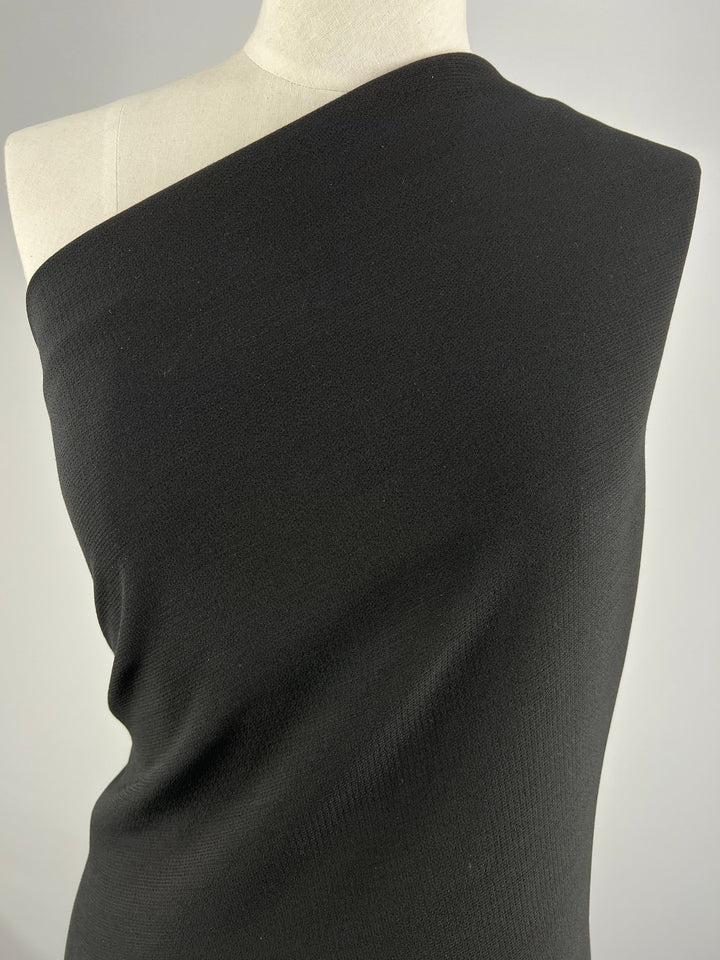 A torso mannequin displays Super Cheap Fabrics’ Plain Crepe - Black - 140cm, styled single-shoulder. The smooth, textured polyester creates a fitted look against a plain light background.