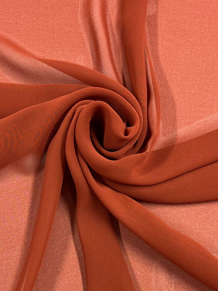 Close-up of a sheer, lightweight polyester fabric in a vibrant terracotta color. The Hi-Multi Chiffon - Picante - 150cm by Super Cheap Fabrics is artfully swirled in the center, creating a spiral pattern that showcases its smooth texture and delicate drape, perfect for floaty tops.