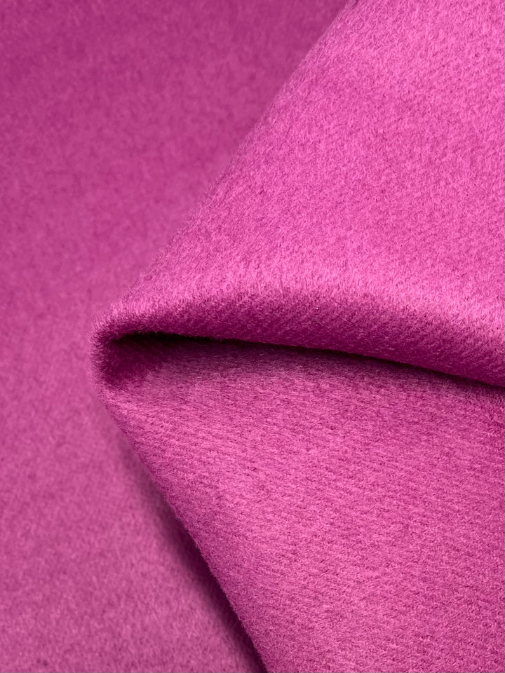 Here's a close-up shot of Super Cheap Fabrics' Wool Cashmere in vibrant raspberry. The 150cm-wide fabric features a smooth, soft texture that is slightly folded at one corner, adding subtle shadows and depth. The fine fibers and rich color highlight the high quality of this material.