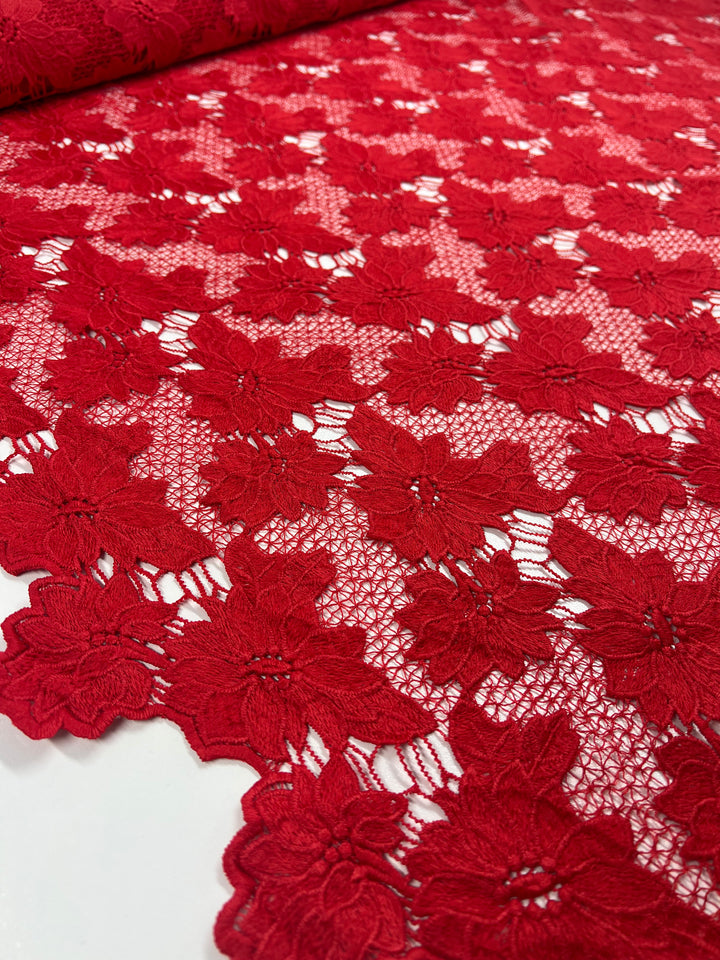 A detailed, close-up image of red floral lace fabric laid out on a flat surface. The heavyweight fabric features intricate flower patterns with an open, net-like design between the petals. The vibrant scarlet color stands out, emphasizing the delicate and elegant texture of the polyester lace. This is Designer Anglaise Lace - Scarlet - 116cm by Super Cheap Fabrics.