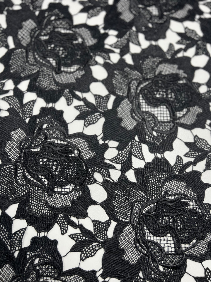 A close-up image of 120cm width black floral lace fabric against a white background. The Designer Anglaise Lace - Black - 120cm from Super Cheap Fabrics features intricate rose patterns and delicate mesh details, showcasing its fine texture and craftsmanship.
