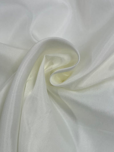 A close-up image of Super Cheap Fabrics' Lining - Cream - 112cm lightweight polyester cream fabric with a subtle sheen. The fabric is gently folded, creating a series of soft creases and curves, highlighting its smooth and silky texture. The folds and the play of light give the fabric a slightly wrinkled appearance, perfect for skirts, jackets, or dresses.