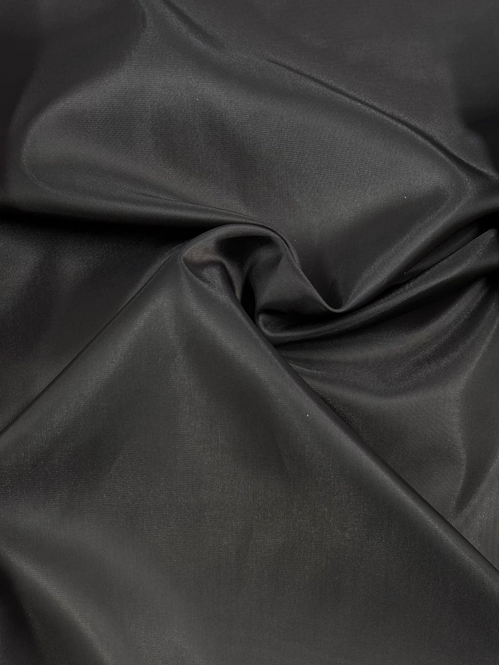 A close-up image of smooth, folded charcoal fabric with a satin-like sheen. This 100% polyester light-weight fabric appears slightly glossy, with soft creases and gentle curves adding texture to the material. The product is Lining - Charcoal - 120cm by Super Cheap Fabrics.