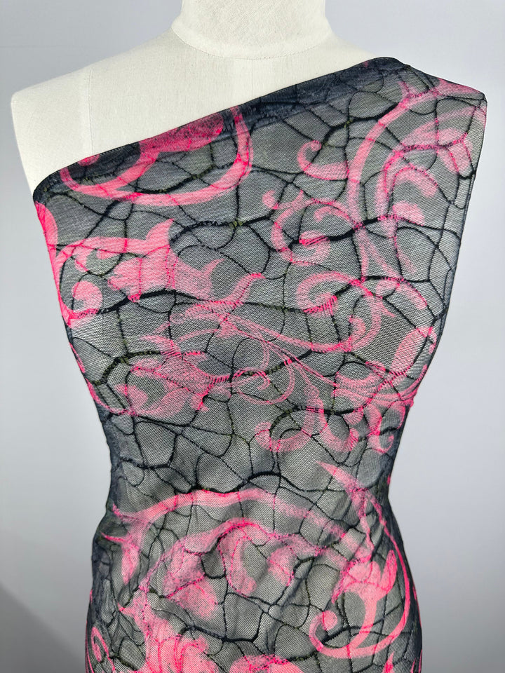 Super Cheap Fabrics' Designer Mesh - Delicate - 150cm is showcased on a mannequin, featuring a lightweight polyester one-shoulder design with black mesh overlay and an intricate pink floral swirl pattern. The white fabric beneath enhances the contrast, offering versatility for any wardrobe.