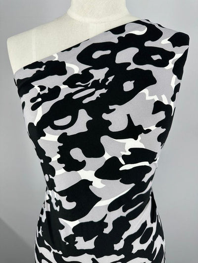 A mannequin displays a garment made from "Printed Lycra - Camo - 150cm" by Super Cheap Fabrics, featuring a one-shoulder design in a black, white, and gray camouflage pattern. The medium-weight fabric is wrapped neatly around the torso, showcasing the asymmetrical design against a neutral background.