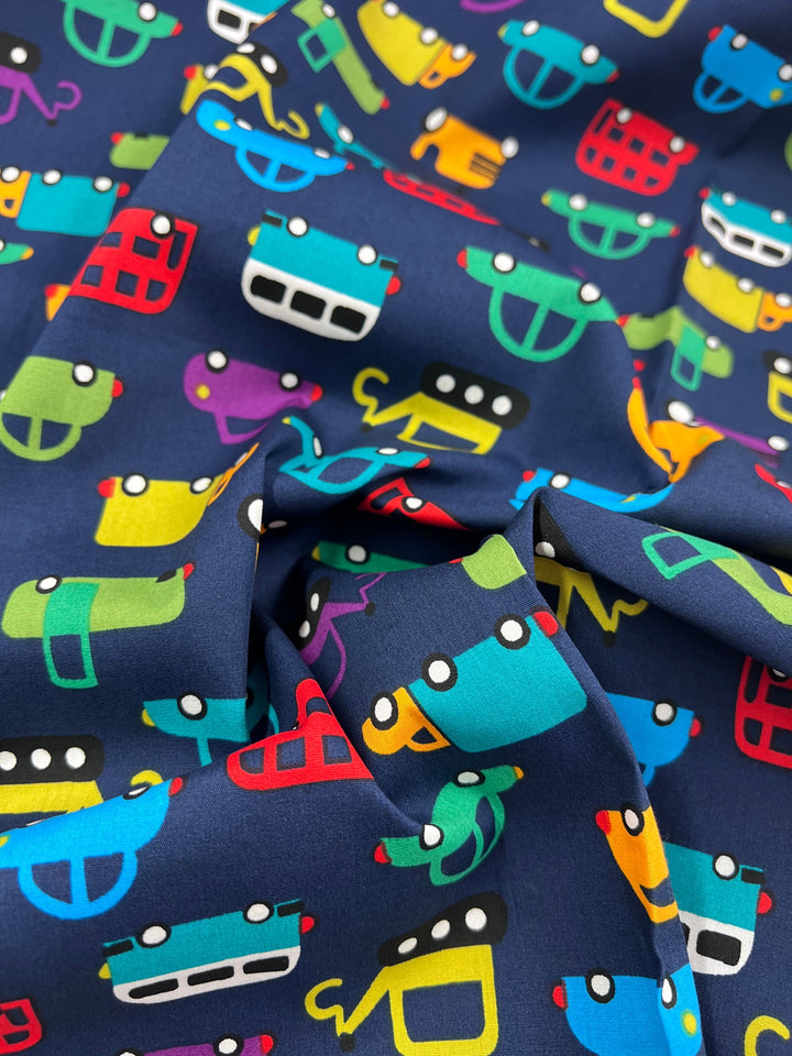 The Super Cheap Fabrics "Printed Cotton - Mobile - 110cm" features cartoon-like vehicles, such as cars and buses, on a dark blue backdrop. Made from natural cotton, the vivid red, yellow, green, and blue vehicles are perfect for quilting or playful decor accents.