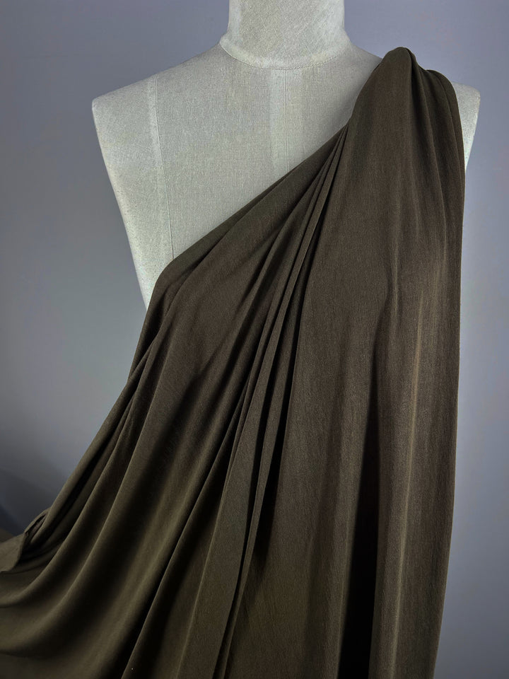 A mannequin wears the Summer Cotton Knit - Sea Turtle from Super Cheap Fabrics, featuring dark brown lightweight cotton fabric that elegantly drapes asymmetrically from the shoulder, showcasing its texture and sophisticated folds against a muted gray background.