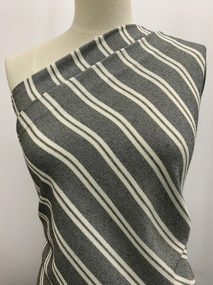 A mannequin is draped with a medium-weight fabric featuring white diagonal stripes on a gray background. This Super Cheap Fabrics blend, Crepe - Zoe Stripe - White, covers one shoulder and angles across the torso, showcasing its texture and pattern against a plain backdrop.