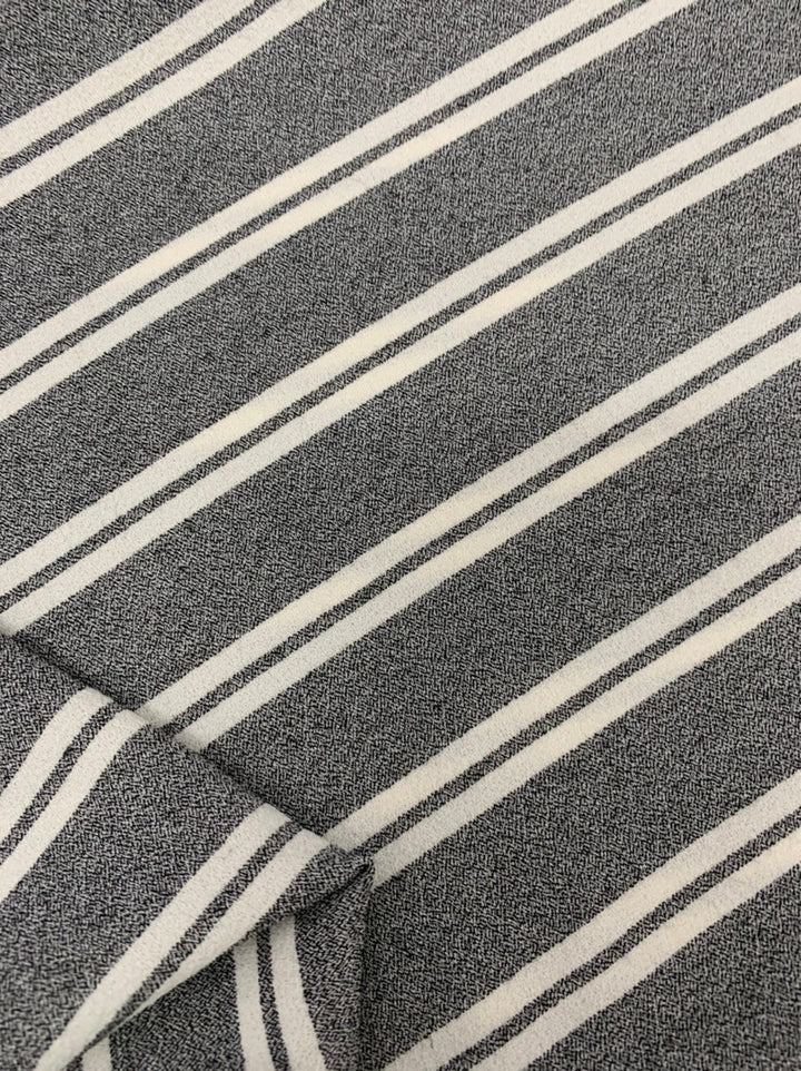 A close-up view of a medium-weight fabric featuring diagonal white stripes on a gray background. The texture of the Crepe - Zoe Stripe - White by Super Cheap Fabrics, made from a polyester viscose elastane blend, is visibly detailed, highlighting the pattern and weave. One corner of the fabric is folded over.