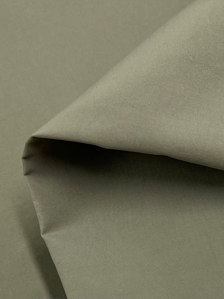 A close-up view of a piece of Delustered Satin - Gray Green - 150cm by Super Cheap Fabrics. The material is slightly folded, showcasing its texture and natural drape. Made from 100% polyester, the lightweight fabric appears to have a matte finish.