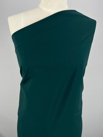 A mannequin draped in a dark green, single-shouldered satin fabric. The 100% Polyester material appears smooth and form-fitting against the torso, creating a sleek and elegant look. The background is plain and light-colored, enhancing the focus on the draped fabric, Delustered Satin - Ponderosa Pine - 150cm by Super Cheap Fabrics.
