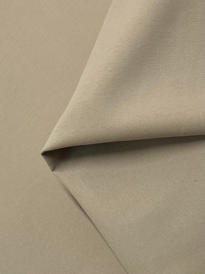 A close-up image of Delustered Satin - Pale Khaki - 150cm by Super Cheap Fabrics, folded diagonally in the center, showcasing its texture and weave. The polyester fabric appears smooth and slightly reflective, with soft shadows accentuating the folds.