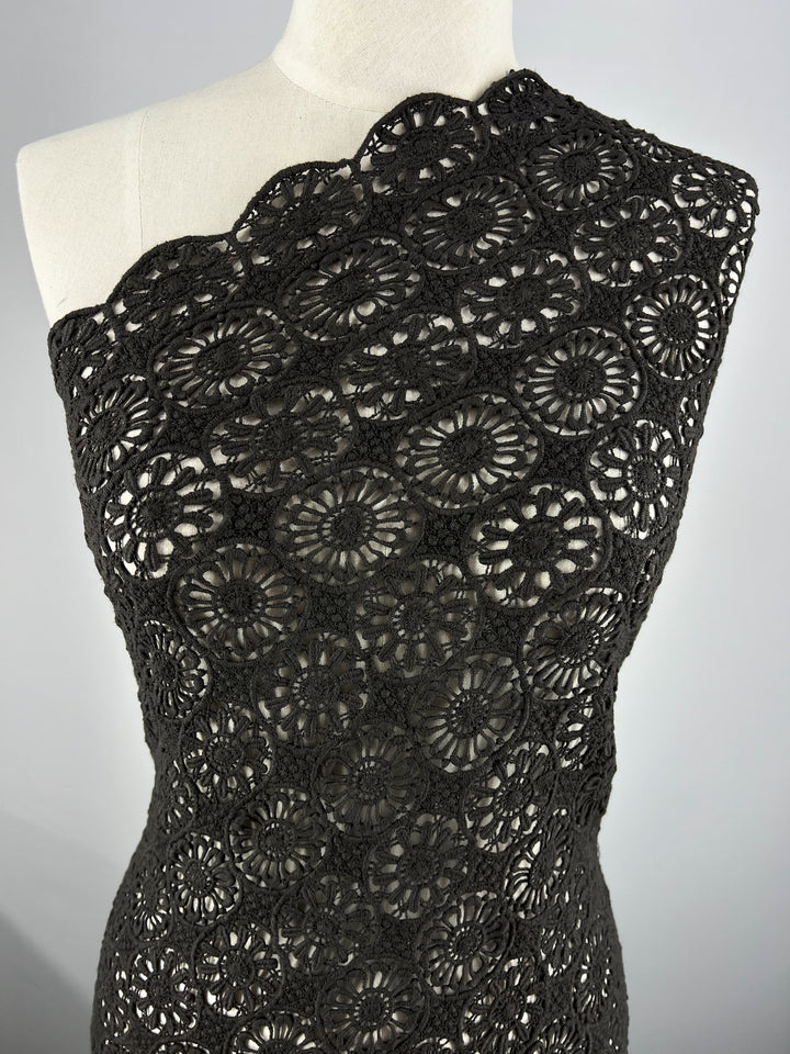 A close-up view of a dress form displaying Anglaise Lace - Black Sun - 110cm from Super Cheap Fabrics. The lace features an intricate floral pattern with various circular and floral designs, making it perfect for bridal wear. The background is a plain light gray.
