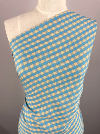 A dress form is draped with Super Cheap Fabrics' Cotton Poly in Cream & Blue Gingham - 150cm, featuring a lightweight gingham pattern in blue and caramel cream. The fabric is styled in a one-shoulder, asymmetrical design, set against a neutral gray background to emphasize the fabric's vibrant pattern and colors.