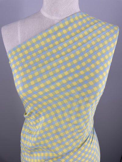 A mannequin is draped in an airy blue and yellow gingham pattern fabric from the Super Cheap Fabrics collection. The Cotton Poly - Blue & Yellow Gingham - 150cm fabric is wrapped asymmetrically around the mannequin, exposing one shoulder. The background is a plain, light gray.