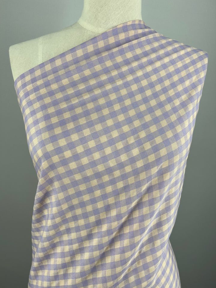 A close-up image of a light weight fabric displayed on a mannequin. The fabric features a small checkered pattern in shades of light purple and beige, reminiscent of a delicate Purple Rose. The background is a plain gray color. The fabric is identified as Cotton Poly - Peach & Purple Gingham - 150cm from Super Cheap Fabrics.