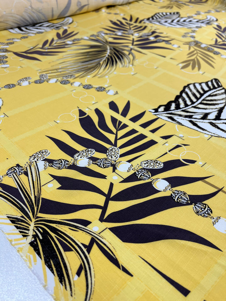 A close-up of vibrant yellow, lightweight fabric, adorned with black and white tropical leaf and botanical designs. The pattern also features a decorative necklace-like chain with beads running across the surface. Perfect for face masks, this slightly crumpled Bamboo Rayon - Pina Colada - 150cm from Super Cheap Fabrics is both functional and stylish.