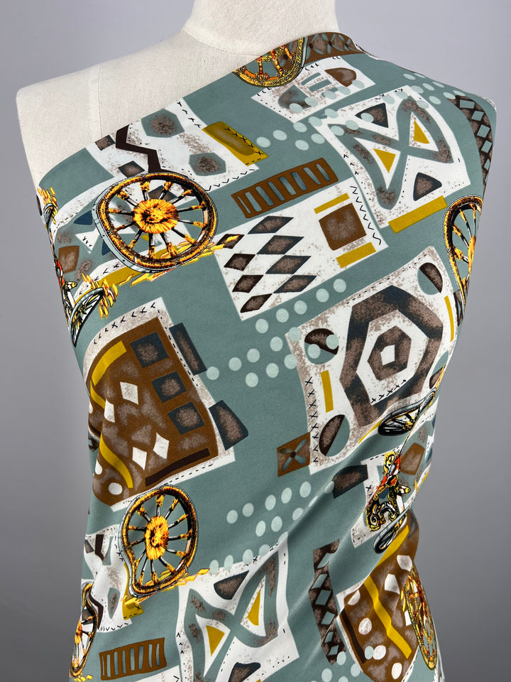 A mannequin draped in Super Cheap Fabrics' Deluxe Print - Medieval - 150cm featuring an abstract design. The pattern includes geometric shapes and intricate motifs in earth tones like brown, white, yellow, and shades of grey on a muted teal background.