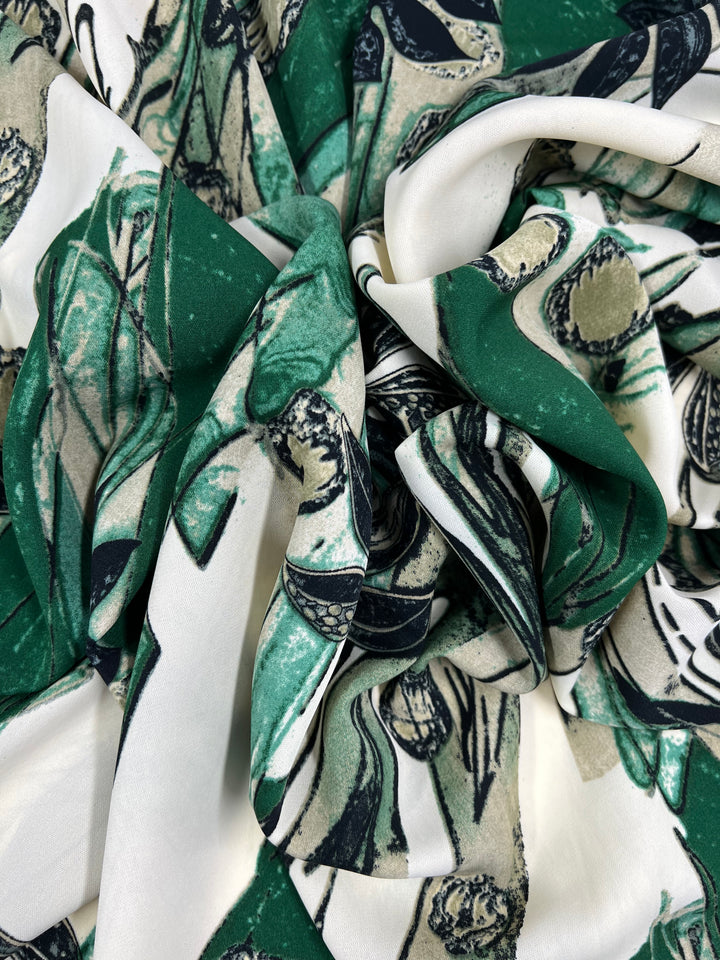 A crumpled piece of versatile polyester with a botanical print featuring shades of green and beige on a white background. The intricate design appears to depict various types of leaves and abstract shapes, giving the easy-care fabric an artistic and natural aesthetic. This is the Deluxe Print - Rebirth - 150cm by Super Cheap Fabrics.