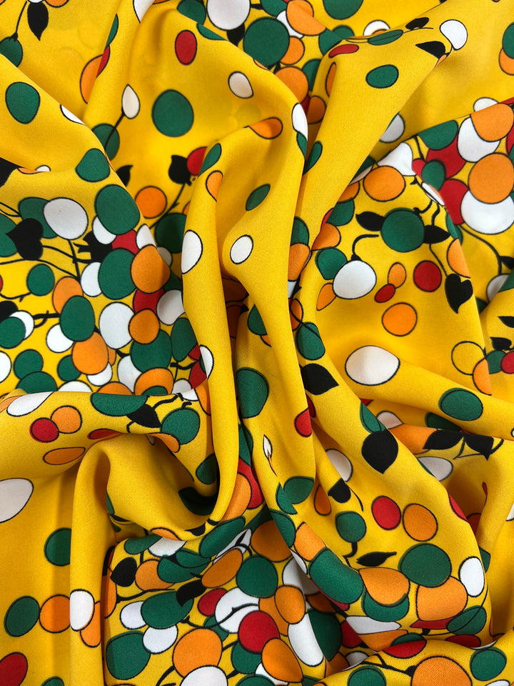 A close-up of a versatile textile with a playful pattern. The fabric is yellow and features small, scattered circles in green, white, black, and orange. Made from polyester fabric, it appears soft and slightly crumpled, creating dynamic folds and shadows. This is the Deluxe Print - Yellow Dots & Vines - 150cm by Super Cheap Fabrics.