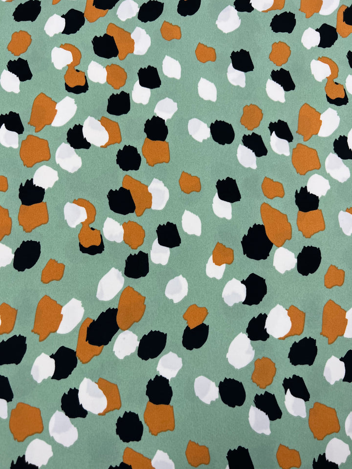 A Deluxe Print - Painters Palette - 150cm from Super Cheap Fabrics with an abstract design featuring irregular spots in black, white, and orange against a light green background. Made from versatile polyester, the spots vary in size and shape, giving a playful and dynamic appearance to this designer fabric.