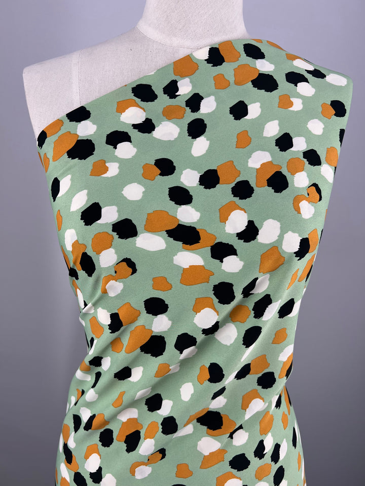 A mannequin is draped with a piece of Super Cheap Fabrics' Deluxe Print - Painters Palette - 150cm, featuring a colorful abstract pattern. The fabric has a mint green background adorned with variously sized spots in black, white, caramel, and mustard colors. The backdrop is plain and softly lit.