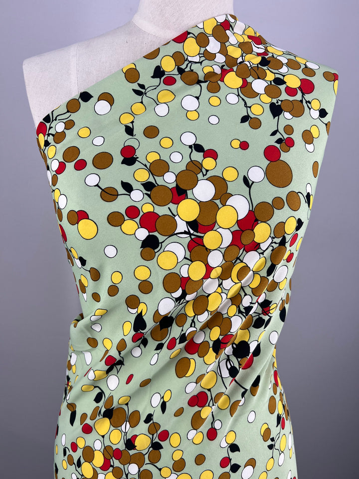 A mannequin is draped with a Deluxe Print - Sienna Dots & Vines - 150cm from Super Cheap Fabrics, featuring a colorful polka dot pattern. The dots are of various sizes in shades of yellow, red, brown, and white, set against a soft green background. The lightweight fabric is wrapped in a way that showcases its pattern dramatically.