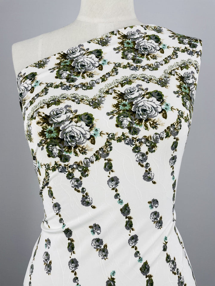 Close-up of a white garment made from medium weight Polyester/Spandex fabric with an intricate floral pattern. The design features large roses in shades of gray, green, and black, arranged in vertical rows and decorative swirls. The fabric is wrapped around a mannequin, showcasing an asymmetrical, one-shoulder style. This is the Printed Lycra - Textured Rose Garden - Grey - 150cm from Super Cheap Fabrics.