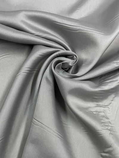 Close-up of a smooth, light gray fabric captured in a swirl pattern. The lightweight fabric has subtle reflections and folds, showcasing its silky texture and elegance. Edges and lines create soft shadows adding a three-dimensional effect to the overall appearance, perfect for garment lining. The fabric featured is Lining - Limestone - 112cm from Super Cheap Fabrics.
