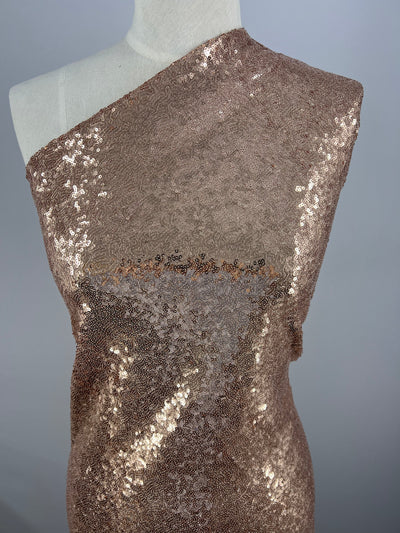 A 150cm width dress form is draped in Super Cheap Fabrics' Evening Sequins - Rose Gold - 150cm, featuring a one-shoulder design. The sequins create a shimmering effect, covering the entire visible surface of the fabric. The backdrop is a plain, light gray color, highlighting the fabric's sparkle.