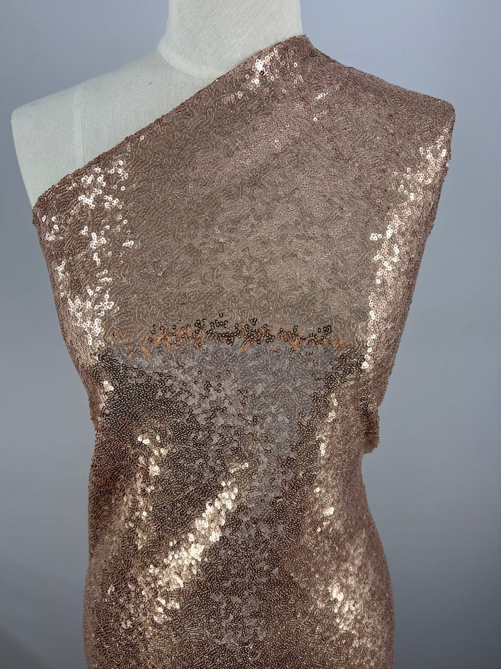 A 150cm width dress form is draped in Super Cheap Fabrics' Evening Sequins - Rose Gold - 150cm, featuring a one-shoulder design. The sequins create a shimmering effect, covering the entire visible surface of the fabric. The backdrop is a plain, light gray color, highlighting the fabric's sparkle.