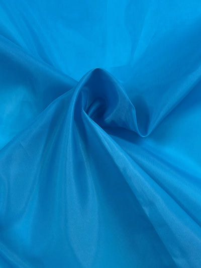 A close-up of a crumpled piece of shiny, vibrant cyan blue fabric. The lightweight lining material has subtle folds and creases, creating a sense of depth and texture. The Lining - Cyan Blue - 150cm by Super Cheap Fabrics appears soft and slightly reflective, catching and playing with light.